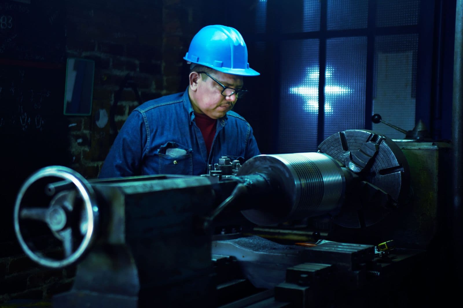 A machinist in a blue hard hat operates a metal lathe in a dimly lit workshop.
